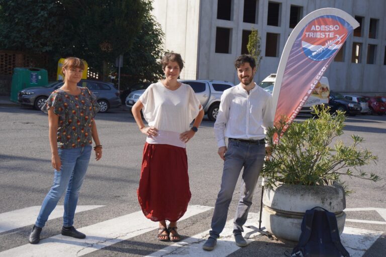 Tre persone sorridenti in piazza assolata davanti a un edificio storico con banner colorato.