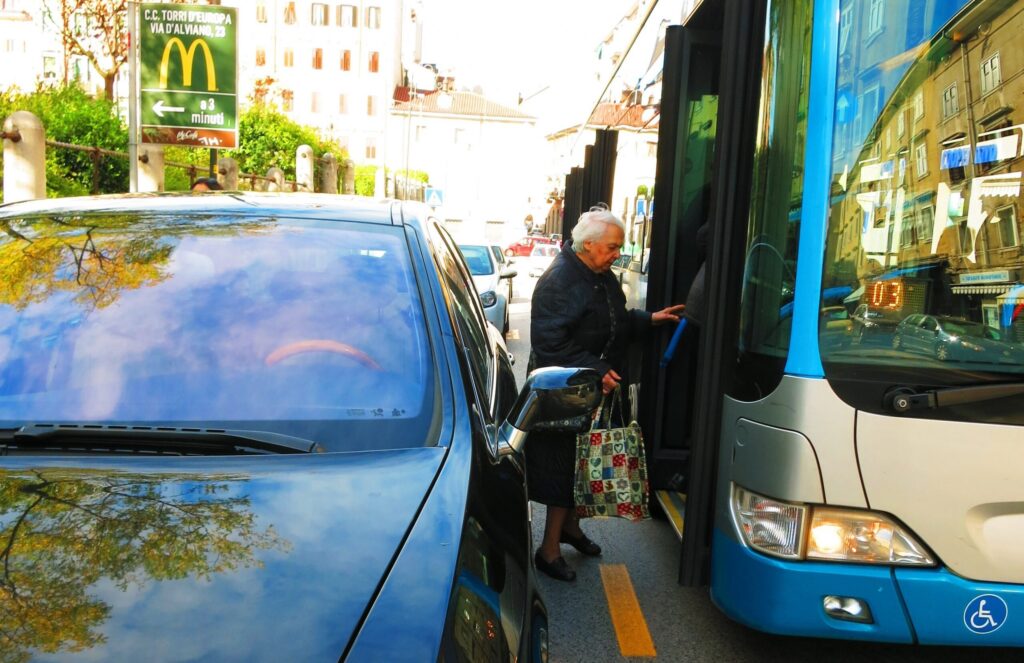 Anziano sale su bus a Trieste tra auto parcheggiata e marciapiede stretto.