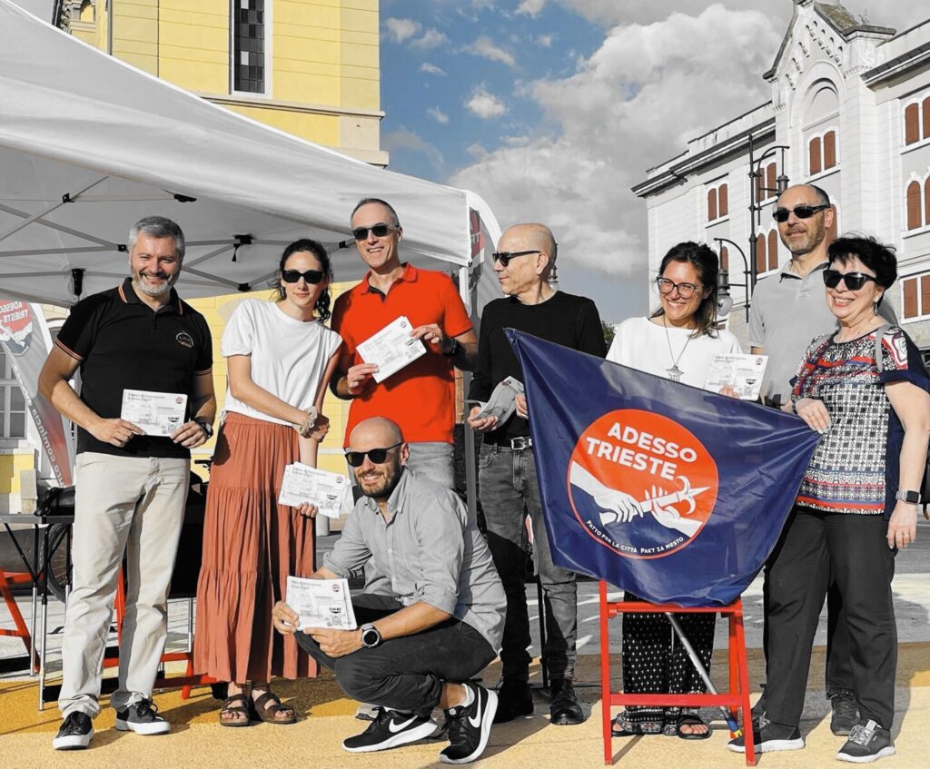 Gruppo Adesso Trieste! in piazza con stand promozionale e cartello “ADESSO TRIESTE!”.