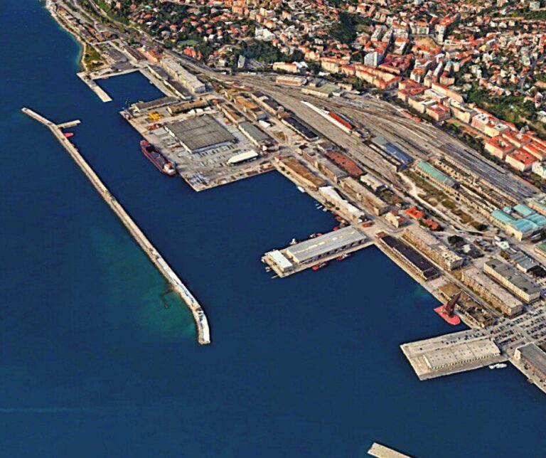 Veduta aerea del porto di Trieste con moli, navi e città storica sullo sfondo.