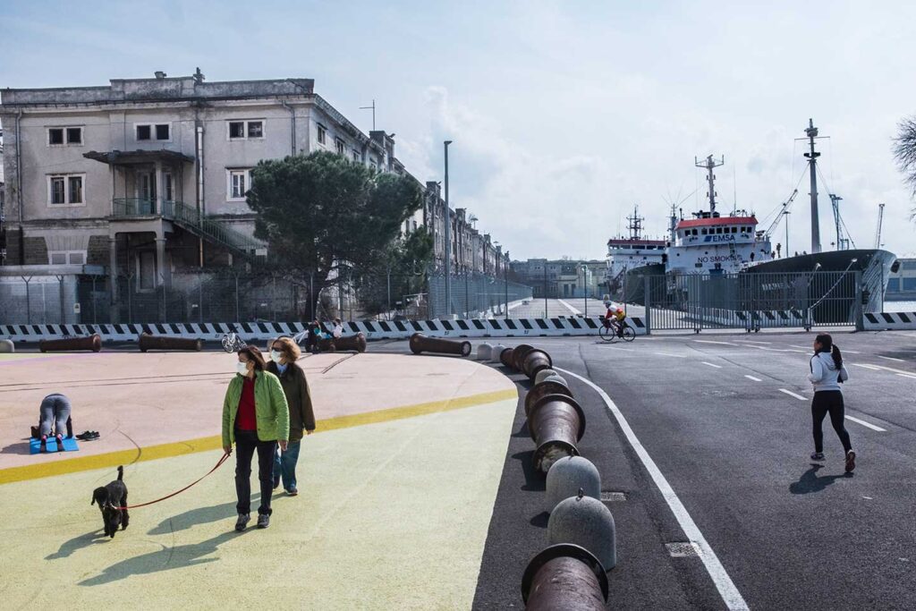 Lungomare di Trieste con passeggiata, cane, palazzi storici e nave in porto.