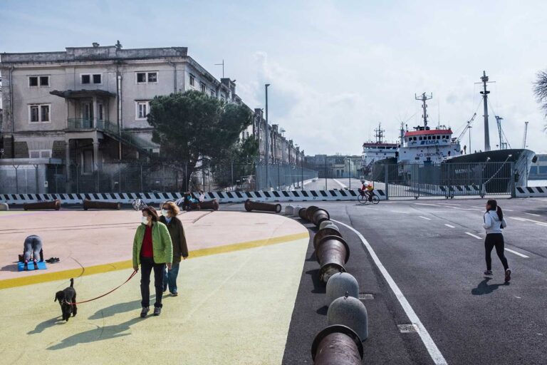 Lungomare di Trieste con passeggiata, cane, palazzi storici e nave in porto.
