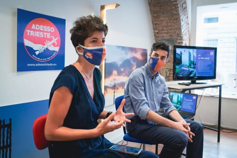 Presentazione ibrida a Trieste con speaker, laptop, monitor e poster ADESSO TRIESTE.