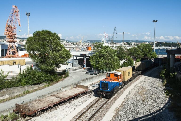 Locomotiva merci nel porto di Trieste con vagoni e gru industriali.