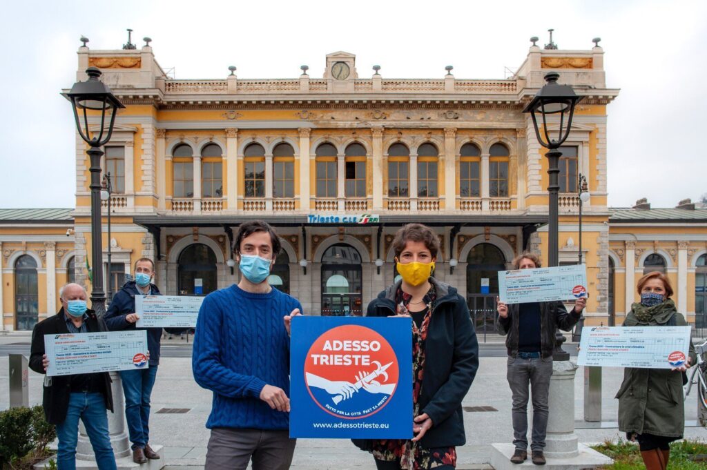 Manifestazione “Adesso Trieste” in piazza storica con manifestanti mascherati e striscione.