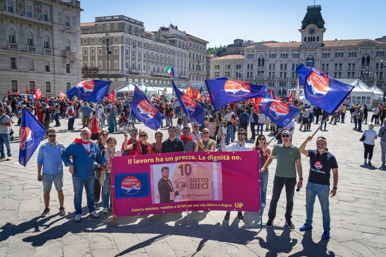 Manifestazione a Trieste con bandiere blu e striscione magenta sull’euro in piazza storica.