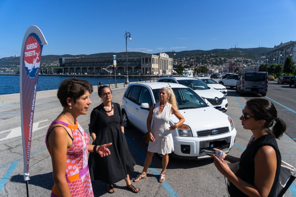 Quattro donne conversano accanto a un’auto bianca sul lungomare di Trieste.