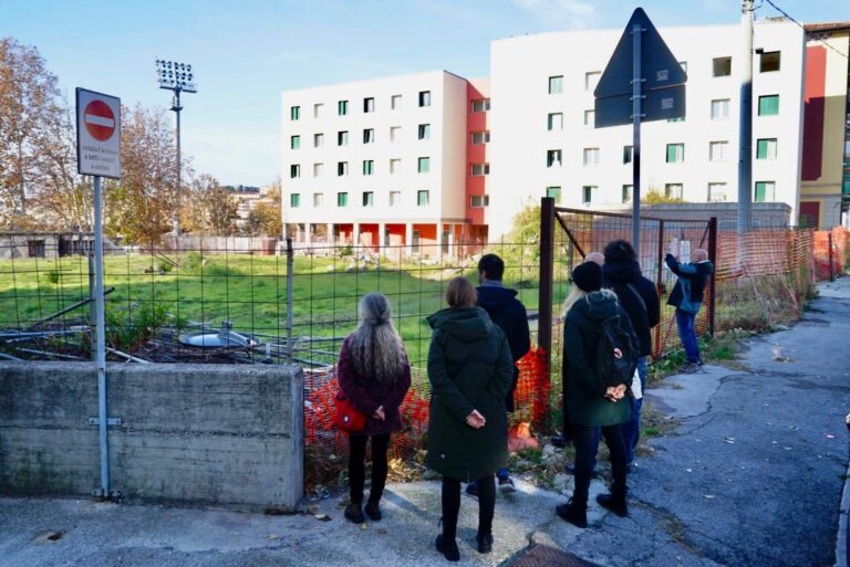Persone in cappotti vicino a una recinzione urbana, con edificio moderno e cielo sereno.