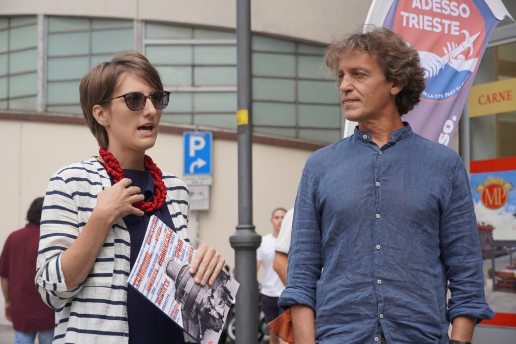 Donna con volantini parla a uomo in camicia blu durante un evento a Trieste.