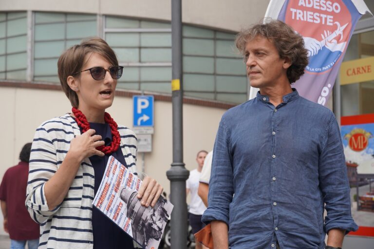 Donna con volantini parla a uomo in camicia blu durante un evento a Trieste.