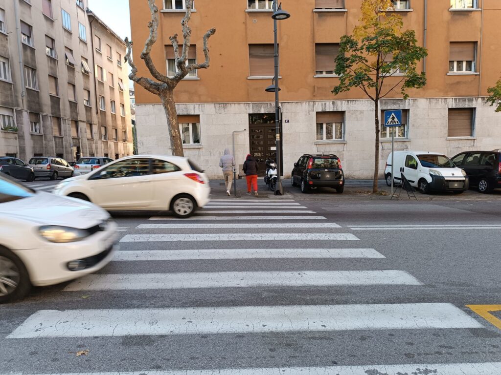 Attraversamento pedonale urbano con auto in movimento e palazzi residenziali sullo sfondo.