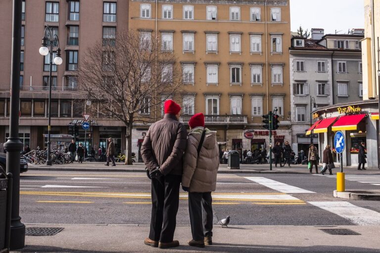 Pedoni in attesa sulle strisce in una strada urbana europea, con piccione e negozi colorati.