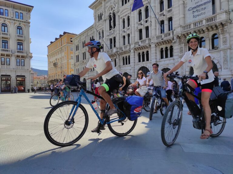 Ciclisti con bici da turismo in piazza storica europea, sotto il sole.