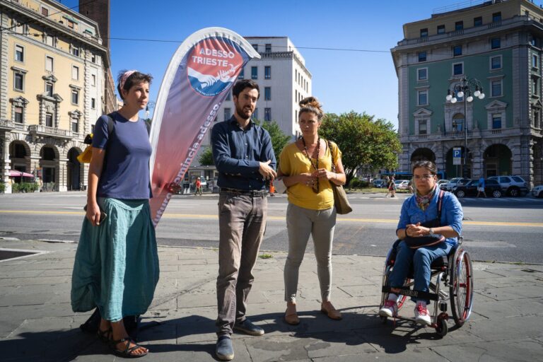 Quattro persone conversano in piazza urbana soleggiata, con una persona in sedia a rotelle.