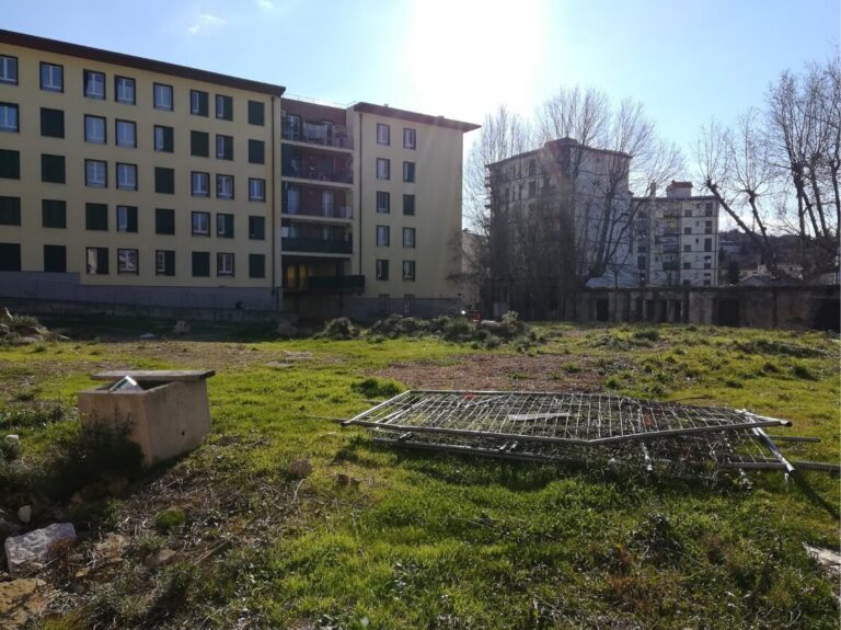 Lotto urbano incolto tra palazzi residenziali, con griglie metalliche e alberi spogli.
