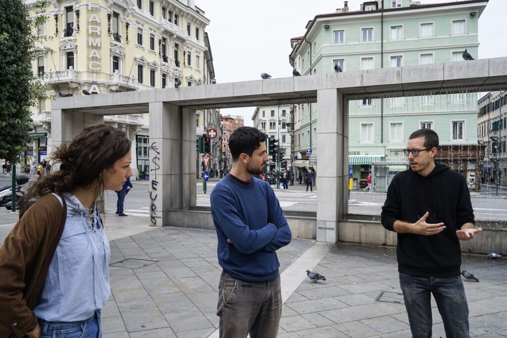 Tre persone conversano in una piazza urbana storica.