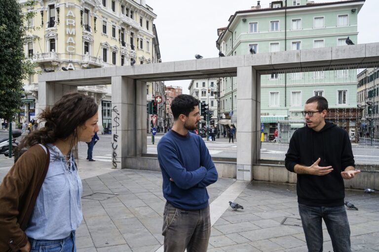 Tre persone conversano in una piazza urbana storica.