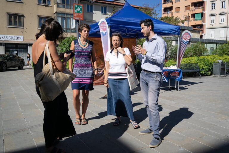 Intervista di gruppo in piazza con microfono, tendone blu e sole.