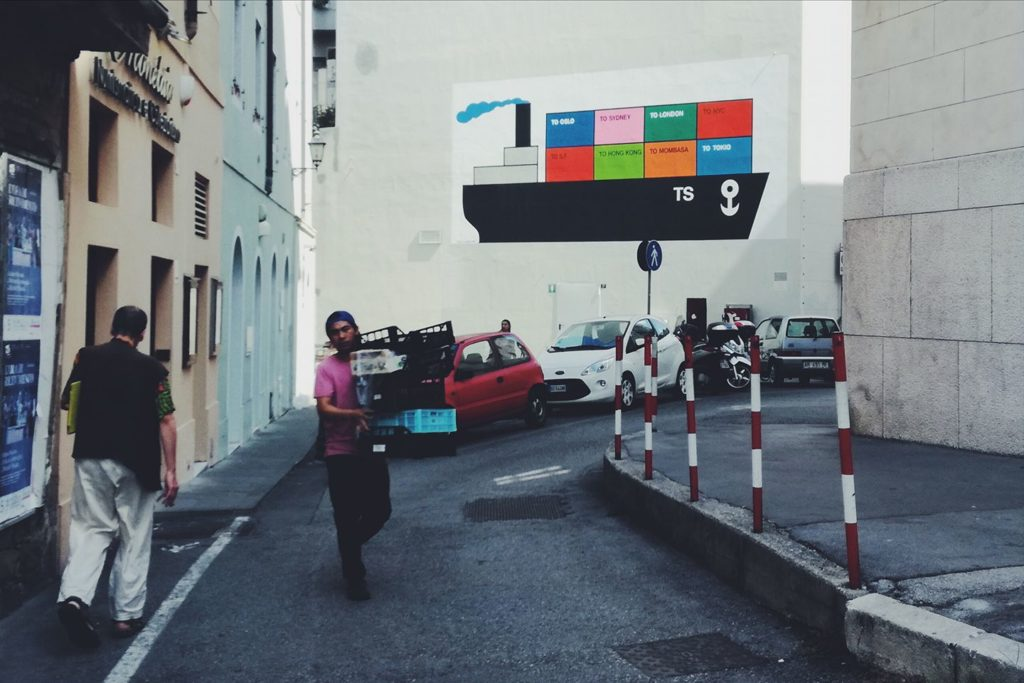 Via stretta con pedoni, auto parcheggiate e murale colorato sullo sfondo.