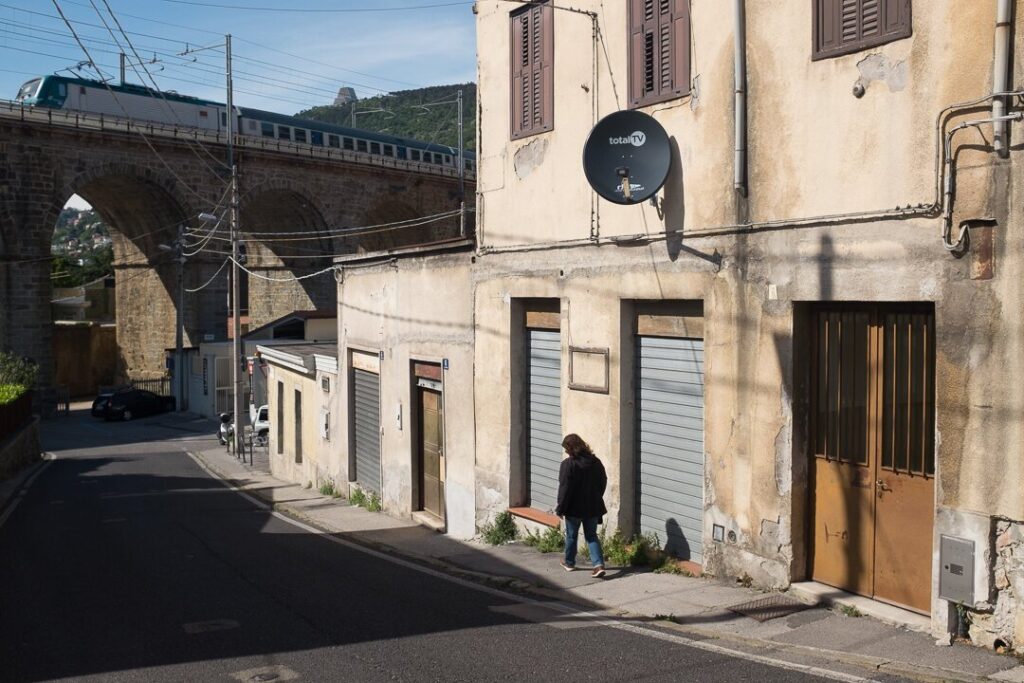 Strada urbana soleggiata sotto viadotto ferroviario storico, con edificio antico e passante.