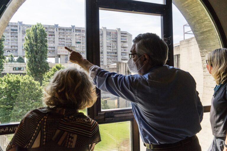 Gruppo osserva il quartiere residenziale dalla finestra, con uomo che indica i palazzi.