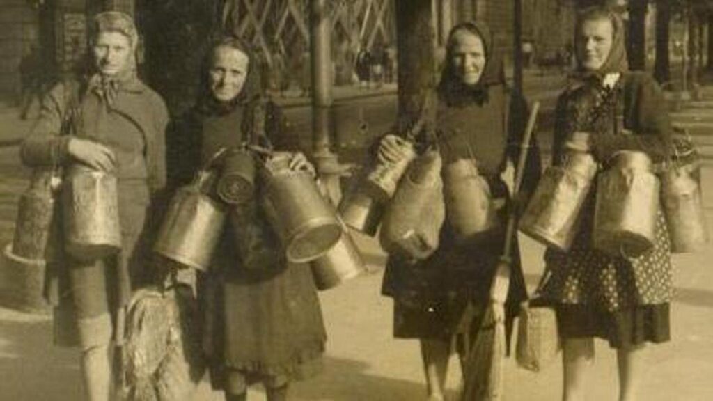 Quattro donne lavoratrici vintage con secchi e attrezzi in una scena storica all’aperto.