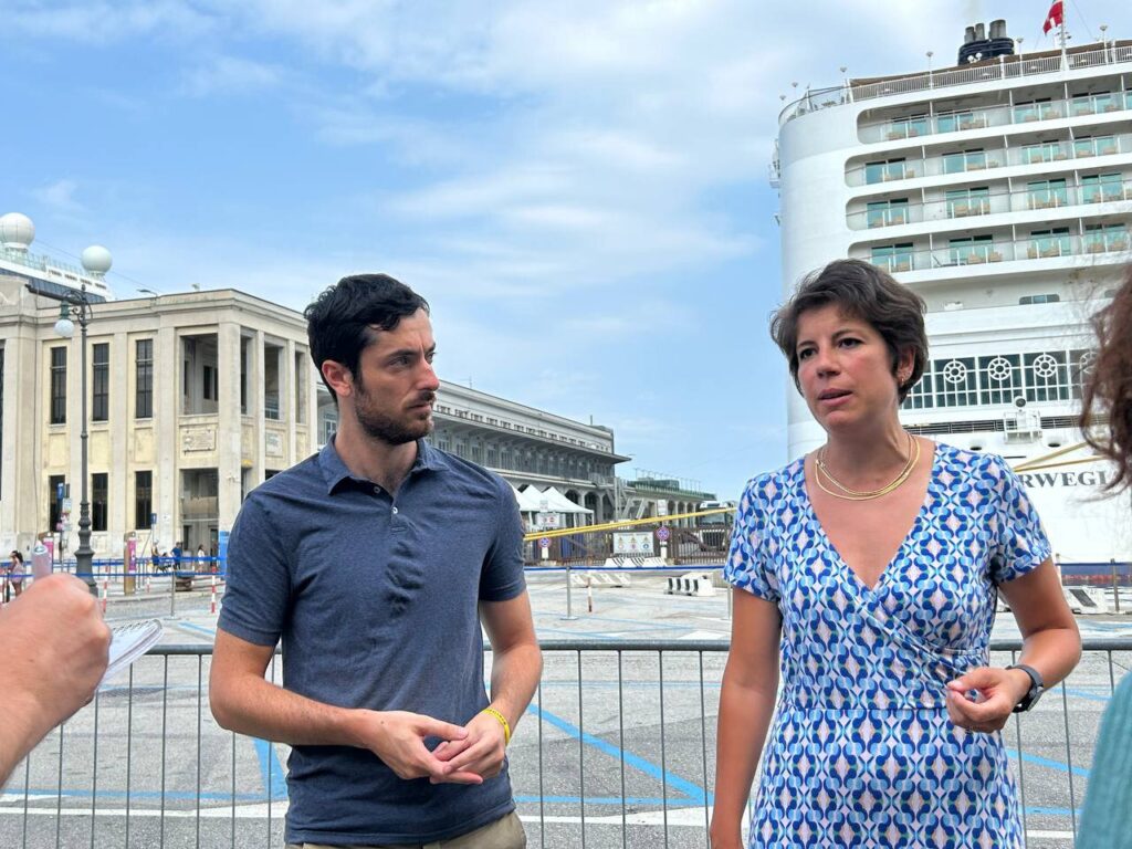 Coppia sul lungomare con nave da crociera e edificio storico sullo sfondo.
