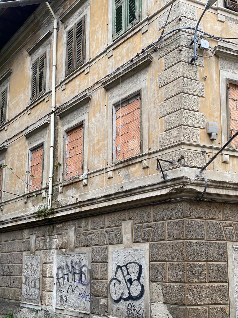Facciata d’angolo degradata con graffiti, pietra a vista e finestre chiuse.