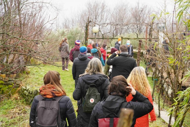 Gruppo in passeggiata invernale nel bosco, tra alberi spogli e sentiero verso una struttura in legno.