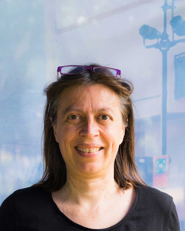 Ritratto di donna sorridente con occhiali, capelli castani e maglia nera su sfondo azzurro.