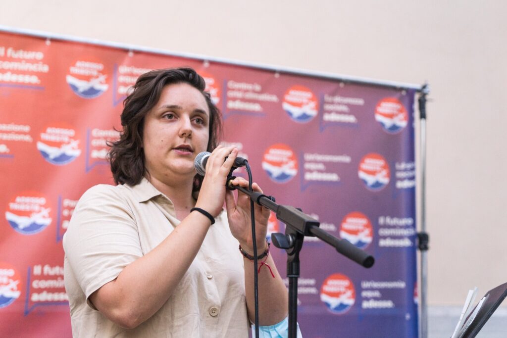 Donna al microfono in un evento ufficiale con sfondo rosso e blu.