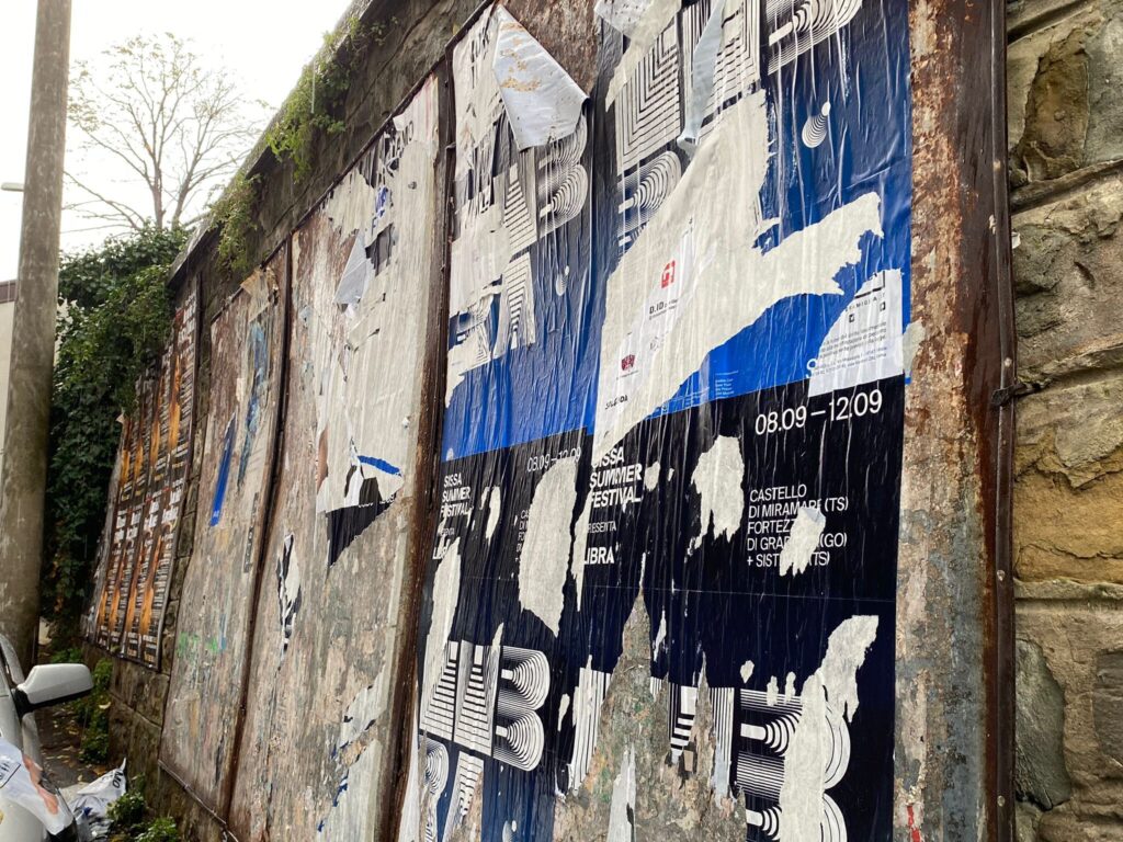 Muro urbano degradato con poster strappati, pietra grezza e scooter in vicolo.